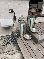 Pair of metal and glass lanterns with white pillar candles inside, two additional white pillar candles on holders, and a tangled set of patio string lights with a remote on the ground next to a wall outlet.