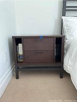 Front and side view of Crate and Barrel wooden nightstand beside bed, showing two drawers with black handles and open shelving compartments, scratches visible on top surface