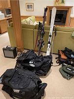 Photo showing all ski equipment including two pairs of skis with poles leaning against couch, ski bags, boots, helmets, and goggles on floor.
