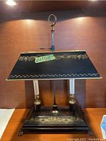 Full front view of the black painted toleware lamp showing two candle bulbs, gold decorative painting, and square base with claw feet.