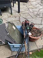 Bin containing two Halloween slate signs, one pot with dried plants, and four tiki torches leaning on the bin and ground on stone patio.
