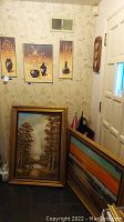 View of multiple framed artworks leaning against wall including floral and garden prints, wooded landscape painting, colorful beach scene, and a wooden thermometer mounted to the wall.