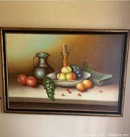 Frontal image of the large still life canvas painting by H.Miller showing fruits, a metal pitcher and candle on table, mounted on wall with frame.