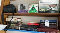 Shelf containing camera bag, boxed binoculars, boxed camera, boxed portable DVD player, and a collection of books and bibles.