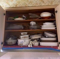 Full view of cabinet with stacked Corning Ware casserole dishes, roasting pans, glass trays, ceramic serving bowls, and kitchen accessories