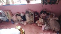 Wide shot of assorted dolls lined against a wall, including boxed dolls, cloth and plastic dolls, some with lace and bonnet outfits, and plush rabbits.