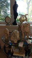 Wide shot showing elephant figurines, small statue, vintage clock, and decorative lyre on glass shelves in front of window.