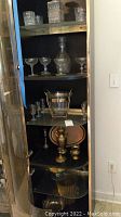 Wide view of shelving with crystal decanters, stemmed coupe glasses, an ice bucket with metal rim, silver platters, and brass metal accessories including small urn and candle holders.