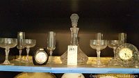 Shelf display showing the decanter with stopper, three etched floral stemware glasses (two coupe glasses and one flute), a small round clock, and a small clear glass vase or bowl.