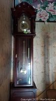 Front view of a tall grandfather clock with carved wooden top, glass door showing pendulum and weights, clock face with Roman numerals, placed against floral wallpapered wall.