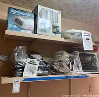 Shelf with boxed air purifier Spring Air 2, boxed clear plastic pitcher set, wrapped health grill deluxe, and various kitchen appliances including an Aroma rotisserie oven, electric skillet, and turkey fryer, plastic-wrapped.
