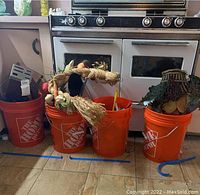 Four orange Home Depot buckets lined up on floor in front of stove, containing various kitchen utensils and decorative items.