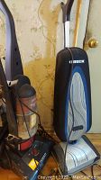 Photo showing two upright vacuum cleaners side by side; Oreck in black with cloth dust bag and Bissell with clear red dustbin.