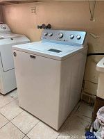 Front and side view of the Maytag washer placed on a tiled floor near plumbing connections.