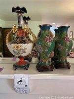 Wide shot showing one porcelain floral urn vase with gilded metal handles and two green cloisonné vases on wooden stands