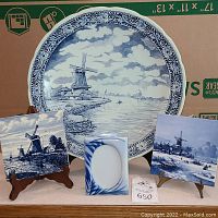 Blue and white Boch decorative plate with windmill and river scene on stand, accompanied by two hand-painted blue and white windmill tiles on small easel stands and a small blue and white leaf-design frame with no visible content.