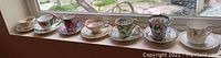 Photo showing all sixteen assorted demitasse cups and saucers displayed in a row on a window ledge, demonstrating the variety in patterns and designs.