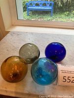 Four glass fishing floats in cobalt, teal, amber, and smoke placed on a white marble surface near window.