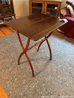 Full view of dark brown wooden TV tray table standing on carpet with visible scratches on top and crossed folding legs with turned stretcher.