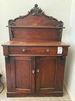 Front view of the vintage sideboard showing one drawer above two cabinet doors, ornate backboard with decorative carving and a shelf. Wood shows some scratches and wear.