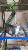 Side view of black and teal green Central Machinery belt sander placed on a metal wire rack, showing the sanding belt and power cord.