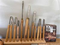 Full view of 10 woodworking chisels with wooden handles in a wooden stand next to a DIY woodworking project book.