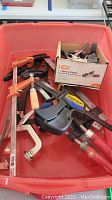 Photo showing assortment of various clamps including quick-grip clamps, spring clamps, and other metal clamps. Clamps have different handle types and colors such as orange, wood, and black.