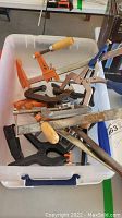 Plastic bin filled with various clamps including several orange bar clamps with wooden handles and metal bars.