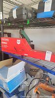 Photo showing Craftsman corded hedge trimmer in red and black box on metal shelving, surrounded by other items including a cardboard box and cases.