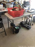 Foldable plastic table with metal legs shown in garage setting, holding various items on its surface and underneath.