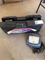 Photo shows black carrying case labeled 'Hydraulic Floor Jack / Gato Hidraulico' and a gray electric charger with power cord placed on floor.