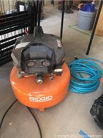 Front angle showing the orange Ridgid air compressor tank and black top housing with connected blue hose.