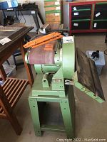 Front side view of green stationary belt sander with sanding belt installed and removable accessories on top, mounted on metal stand