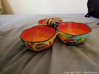 Front and side views of the tri-sectional clay dish showing the bright orange interior, yellow and multicolored exterior with chili pepper and Mexican hat motifs, and the central chili pepper-shaped handle.