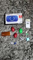 Full lot display showing Monster Superstar speaker box, three egg shakers (red and green), two iPod Shuffles (black and blue), Jackery battery charger (orange), two guitar picks, white USB charging cable on carpet.
