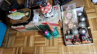 Assorted Christmas decorations including novelty ornaments shaped like fish and trains, glass baubles and floral glass ornaments, ceramic lighthouse decoration, all placed in two cardboard boxes on a wooden floor.