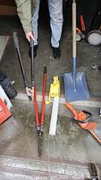 Photo showing manual garden loppers, hedge shears, shovel, and black and orange electric hedger and line edger tool.