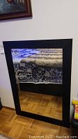 Full view of rectangular wall mirror with black frame hanging against a wall. Floor and part of a blanket are reflected in the mirror.