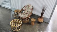 Overall lot showing one rattan swivel rocker chair with floral cushion, a rattan ottoman or table, a small plant stand, and decorative woven vase with dried branches.