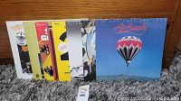 Eight vinyl record albums standing on a carpet against a wooden wall, with Air Supply album at front showing blue cover with hot air balloon.
