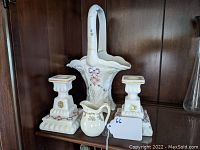 Photo showing two square-based milk glass candlesticks decorated with purple bows and floral accents, a tall milk glass basket with floral decoration and a repaired handle, and a small cream pitcher with gold floral pattern