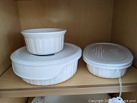 Three white CorningWare French White pieces arranged side by side, including oval dish, round lidded dish, and smaller lidded ramekin on the right.