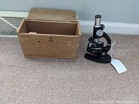 Black Atco BR2 microscope beside open original wood storage case on carpeted floor