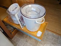 Electric ice cream maker base in white plastic with transparent lid, additional removable parts wrapped in plastic, and booklet placed beside it on wooden surface.