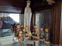 Shelf display of the group of Hummel and Goebel porcelain figurines including a religious figure and several children figurines.