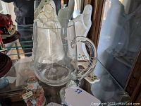 Photo showing clear etched glass beer stein in display cabinet with porcelain figurines behind it
