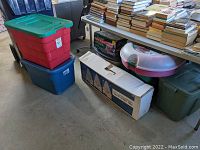 View showing multiple plastic storage bins stacked on floor and various boxes under a table with books on top, containing Christmas decorations.