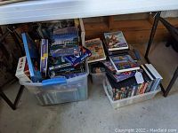 Large quantity of DVDs in two plastic bins under a table. The DVDs are stacked and disorganized showing various cover art.