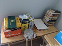 Photo showing stacked manuals, books, some boxes, a metal tin, and an aluminum pot with lid on a wooden table.