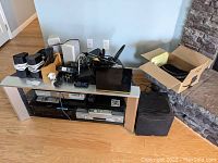 Photo showing RCA surround sound speakers, subwoofer, Blu-ray and DVD players on a glass shelf TV stand with remote controls and cables spread on top.